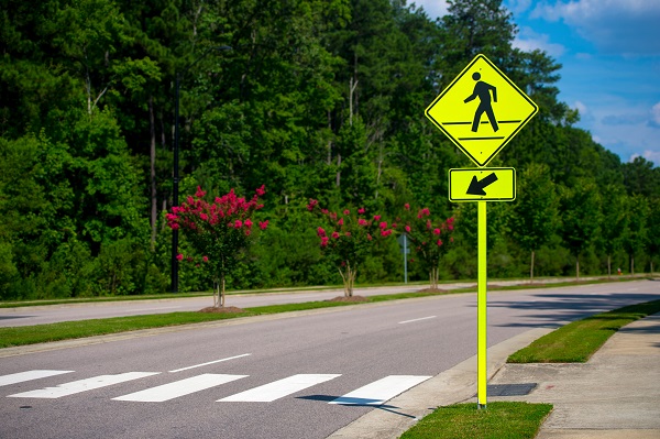 Pedestrian crossing sign with an arrow pointing to a crosswalk on a tree-lined road, with vibrant green trees and pink flowering bushes in the background.