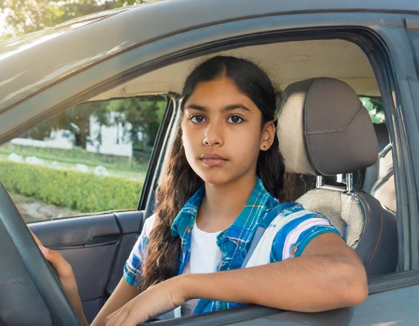 A teenage girl with long dark hair is sitting in the driver's seat of a car, looking directly at the camera with a serious expression. She is wearing a blue plaid shirt over a white t-shirt. Her right hand is on the steering wheel, and her left arm is resting on the car door. The car window is rolled down, and the background shows a sunny day with green bushes and trees.